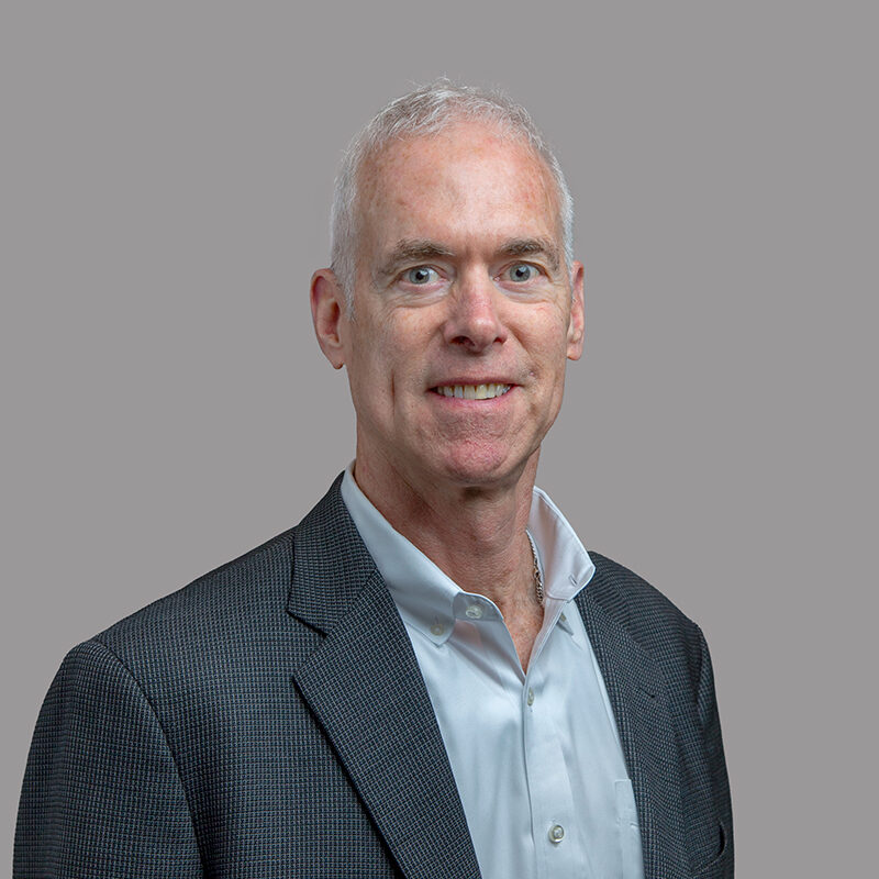 Bill Colligan smiles neutrally at the camera. His gray blazer and blue shirt complement his features.